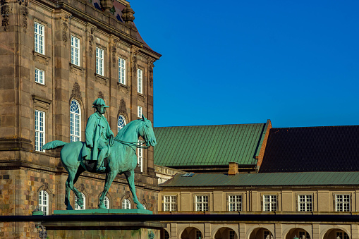 Christiansborg Palace with the statue of King Christian IX, the statue was erected 1906, and a clear polarized sky behind. In the foreground is the square in front of the palace which is a government building. Christiansborg Palace is a palace and government building on the islet of Slotsholmen in central Copenhagen, Denmark. It is the seat of the Danish Parliament, the Danish Prime Minister's Office and the Supreme Court of Denmark.