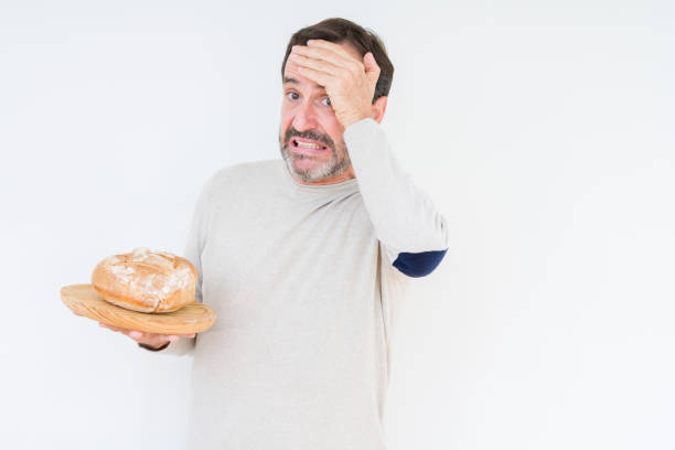 istock Senior man holding homemade fresh bread over isolated background stressed with hand on head, shocked with shame and surprise face, angry and frustrated. Fear and upset for mistake. 1084171502