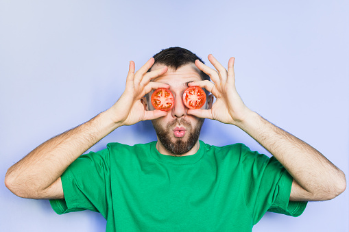 Young bearded man holding slices of red tomatoes in front of his eyes, surprised face expression. Light purple background, copy space.