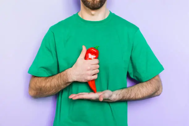 Young man holding red capi pepper in his hands showing thumb up on green t-shirt background. Light purple wall on background, copy space.