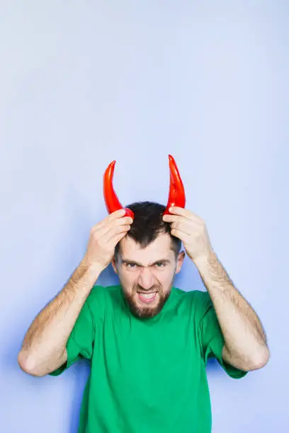 Young man holding red capi peppers above his head which looks like horns, angry and evil face. Light purple background, vertical orientation, copy space.