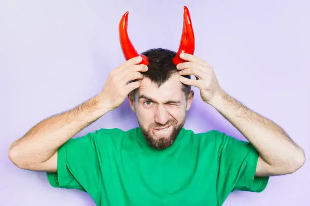 Young man holding red capi peppers above his head which looks like horns, angry and evil face. Light purple background.
