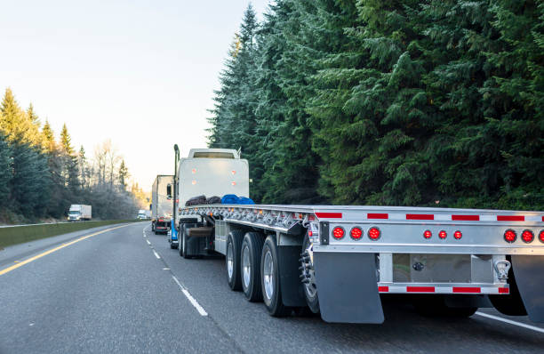 Big rig long haul semi truck with empty flat bed semi trailer running on the winter road in convoy with another semi trucks Big rig long haul powerful semi truck with empty light weight aluminum flat bed semi trailer running on the winter road in convoy with another semi trucks and evergreen trees trailer stock pictures, royalty-free photos & images