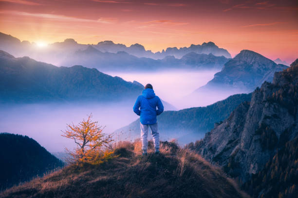 istock Man on the mountain peak looking on mountain valley with low clouds at colorful sunset in autumn in Dolomites. Landscape with traveler, foggy hills, forest in fall, beautiful sky at dusk in Alps 1084242964