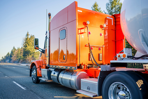 Bright orange big rig American professional long haul semi truck transporting tank semi trailer for transportation of liquid and liquefied chemical commercial cargo running on the road