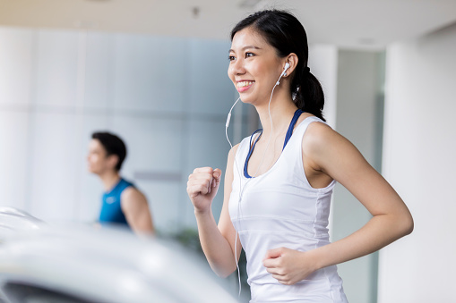 In this side view, a smiling young woman looks straight ahead as she runs on a treadmill at her health club.  She is listening to music on a pair of earbuds.