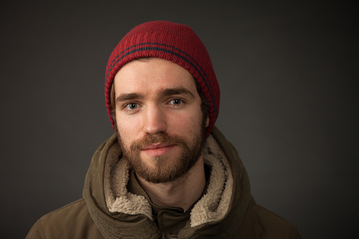 Studio portrait of a 25 year old bearded man in a knitted hat and winter coat on a black background