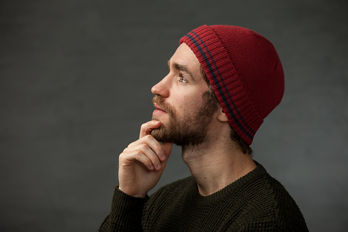 Studio portrait of a 25-year-old bearded man in a knitted hat and sweater on a gray background