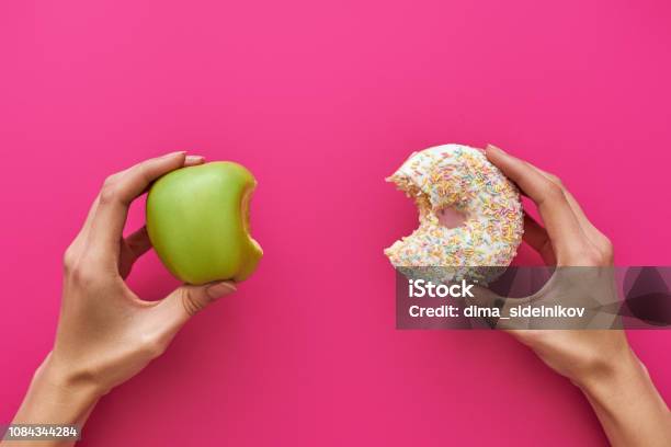 Dieting Or Good Health Concept Young Woman Trying To Choose Between Apple And Donut Stock Photo - Download Image Now