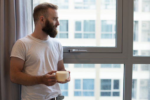 Serious pensive young bearded man drinking coffee and looking out window. Handsome guy with cup standing at window at home. Contemplating concept