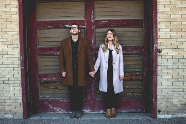 lindo largo humorístico retrato de una pareja hipster posando delante de una antigua puerta de garaje - couple old fashioned hipster holding hands fotografías e imágenes de stock