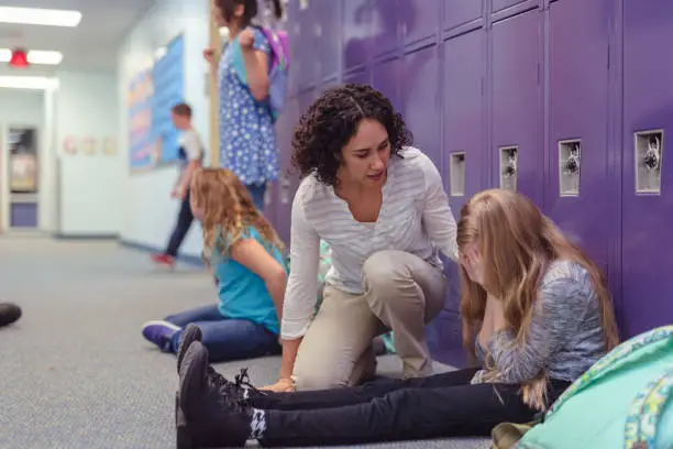 A young left-out elementary student sits by her locker with her face covered in hands while others leave her out and play with each other. An ethnic female teacher is crouching down to console her and find out if she's okay.