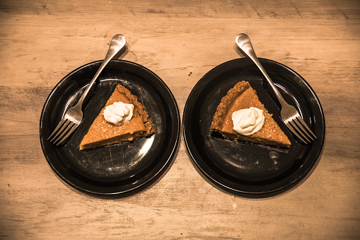 Two fresh slices of pumpkin pie on black plates with whipped cream on top and forks. Viewed from above, sitting on wood table. Bright. Seeing double. Whole wheat crust