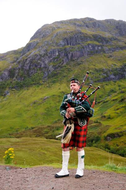 piper en el tradicional traje escoc toca gaita en tierras altas de escocia en el fondo de la montaña. nublado día de otoño. - bagpipe fotografías e imágenes de stock