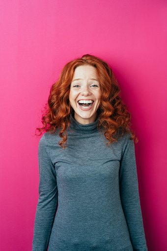 Cute young redhead woman laughing at the camera as she poses over a bright pink studio background with copy space above