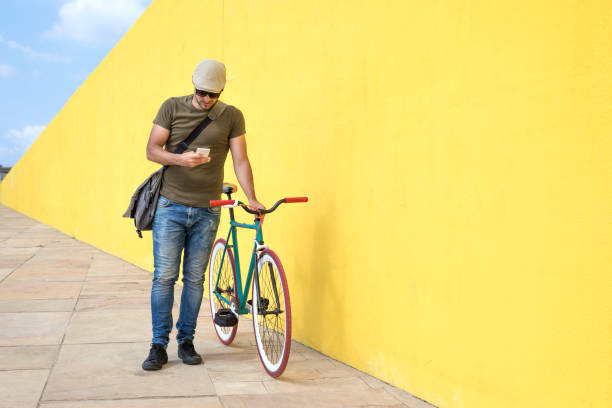 istock Full length of man using smart phone while standing by bicycle against yellow wall at sidewalk in city 1084440028