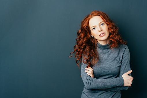 Pretty young redhead woman staring intently at the camera with folded arms and parted lips over a dark studio background with copy space