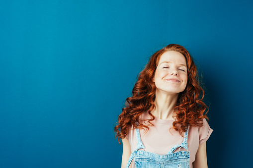 Blissful young redhead woman with a happy smile looking up to the side with closed eyes over a dark studio background with copy space