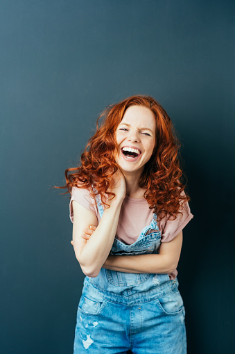 Happy vivacious young woman in trendy designer dungarees standing with her hand to her neck having a good laugh over a dark studio background with copy space