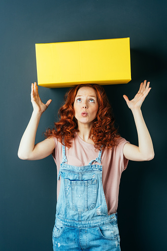 Young red-haired woman wearing denim overalls with yellow box on her head