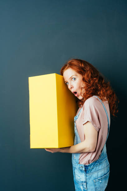 istock Young cheerful red-haired woman with yellow box 1084451764