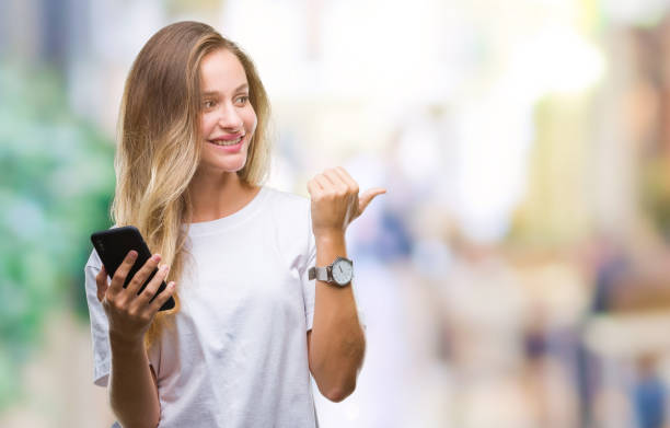 istock Young beautiful blonde woman using smartphone over isolated background pointing and showing with thumb up to the side with happy face smiling 1084454936