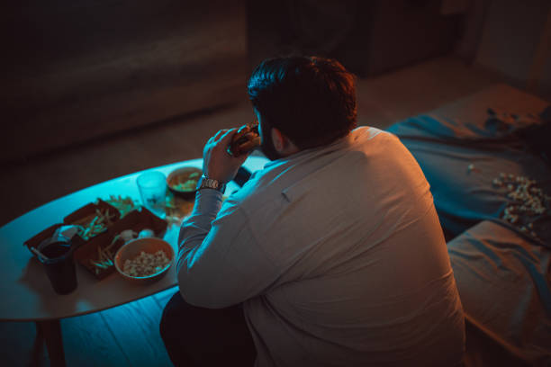 Overweight man eating a burger One man, sitting at home, eating burgers and watching a movie alone, rear view. evening meal stock pictures, royalty-free photos & images