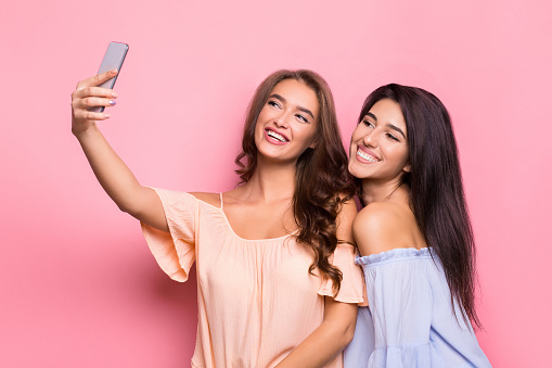 Selfie time. Two smiling women posing at smartphone camera, pink background
