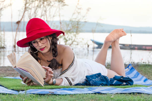 Portrait of young beautiful woman wearing pale pink dress reading book against background of pond water in park on summer day. Reading book in nature. Back view.