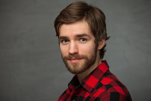 Studio portrait of a 25 year old bearded man in a red plaid shirt on a gray background