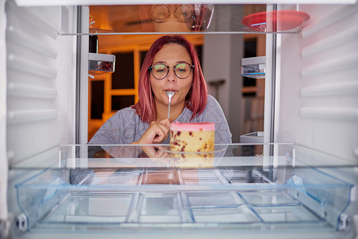 Woman eating gateau while standing in front of fridge. Picture taken from the inside of fridge.