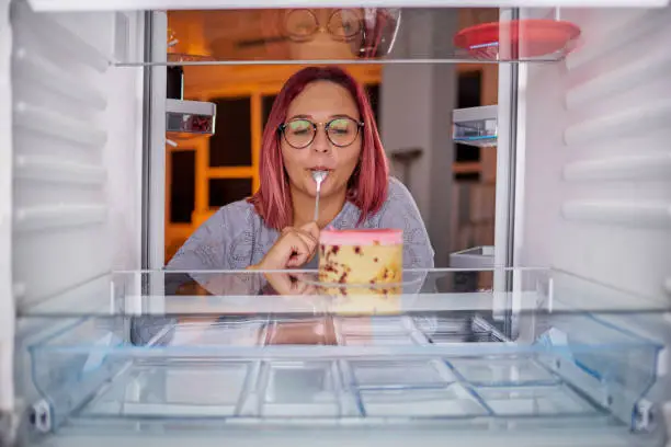 Photo of Woman eating gateau while standing in front of fridge.