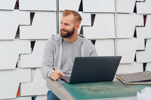 Young bearded man sitting at the desk with laptop against grey decorated background and looking away with a smile