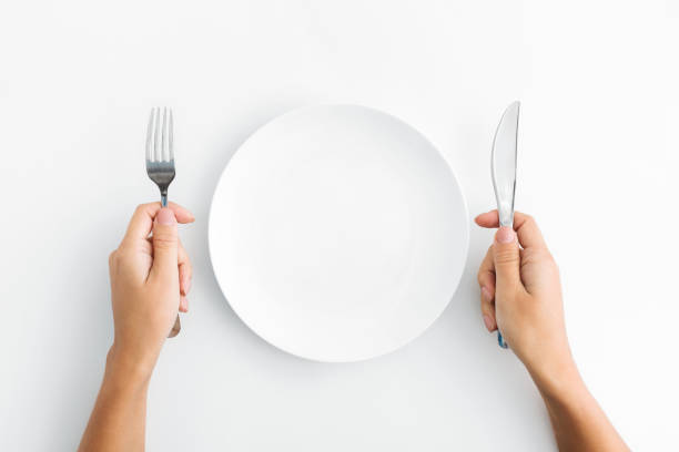 Whte plate with silver fork and knife on white background Dinner place setting. White empty plate and hand holding silver fork and spoon on white background, top view, copy space empty plate stock pictures, royalty-free photos & images