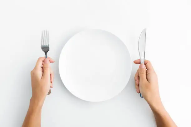 Dinner place setting. White empty plate and hand holding silver fork and spoon on white background, top view, copy space