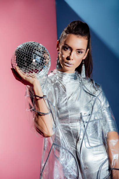 istock beautiful model in silver bodysuit and raincoat posing with disco ball on pink and blue background 1084551056