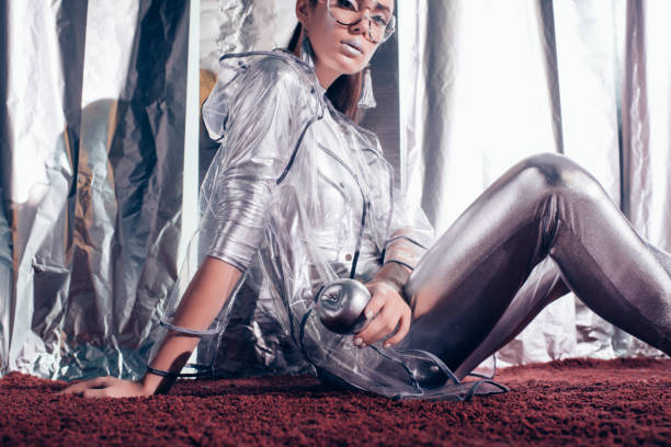 elegante mujer joven en traje de plata y capa impermeable sosteniendo manzana plata y posando para sesión de moda sobre fondo metálico - macintosh apples fotos fotografías e imágenes de stock