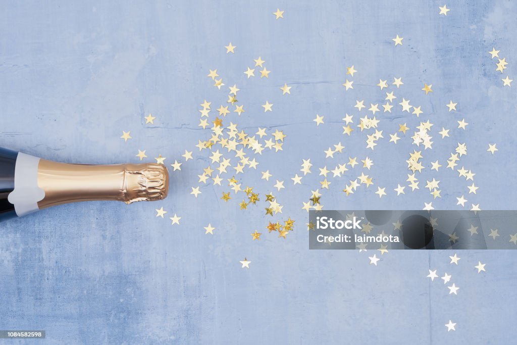 Champagne bottle with confetti stars on blue background. Holiday concept. Flat lay style. Copy space, top view. Champagne bottle with confetti stars on blue background. Christmas, birthday or wedding concept. Flat lay style.  Copy space, top view. Champagne Stock Photo