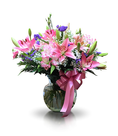 A large fresh vibrant multi colored flower arrangement of pink lily blossoms and buds, white gypsophila, green leaves and a small purple flower cut out with a white background. This beautiful bouquet is decorated with a bright pink ribbon tied around the clear glass vase. There is a faint reflection on the table surface.