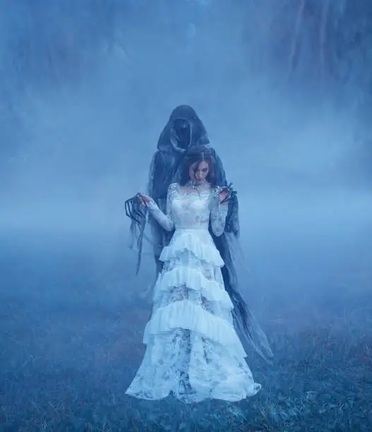 Photo of Dark Lord's Bride in white vintage mesh translucent dress and a silver necklace is standing on the frozen grass in a thick blue fog in monster hands. Halloween costume idea. art photo.