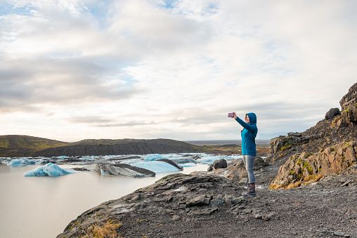 This photograph is of an American woman in her 30s taking pictures with her mobile phone while hiking along a glacier lagoon in Southeastern Iceland.
