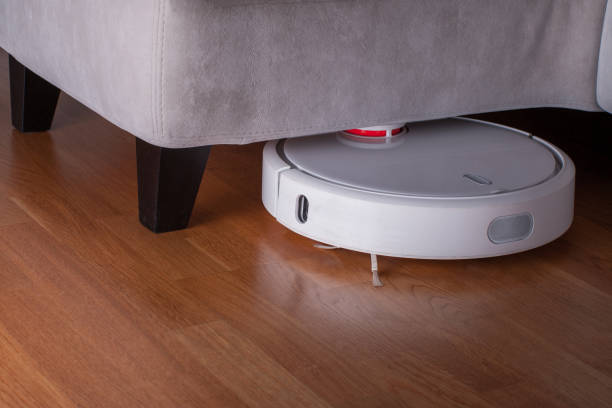 istock white Robot vacuum cleaner runs in corner under sofa on wood parquet floor. Modern smart cleaning technology housekeeping 1084705314