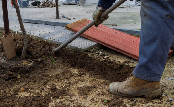 Worker digging a ditch with a hoe in a construction in a rural place. Man in boots and safety gloves. Many construction materials in the background.