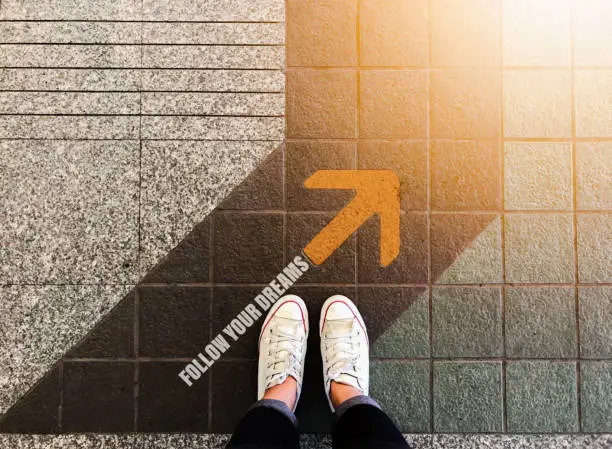 Photo of Follow your dreams yellow arrow and woman feet wear white sneakers shoe on floor background