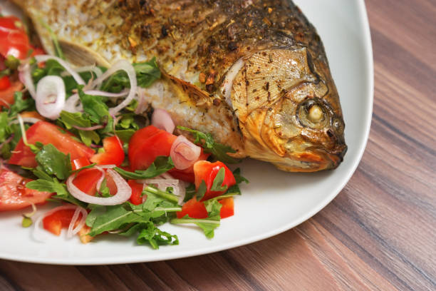 istock Fish dish. Roasted crucian carp on white plate with fresh mix vegetable on table. Close-up fried carassius ready-to-eat. Hot dish. Main course. 1084824162