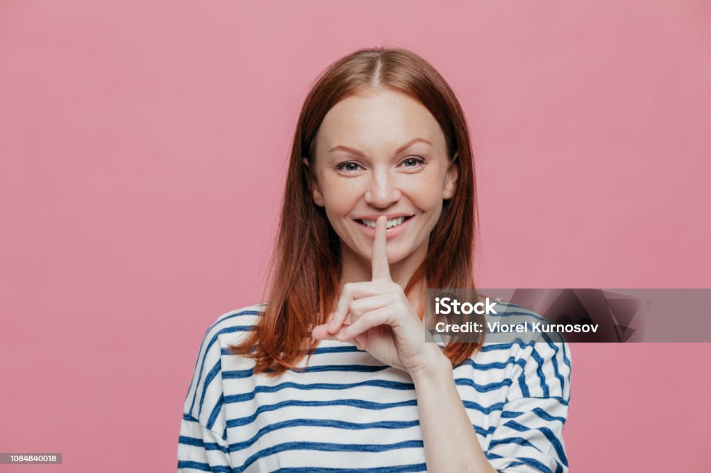 Pleased attractive woman with brown hair, gentle smile, keeps index finger on lips, shows silence gesture, dressed in striped clothes, models over pink background. People and conspiracy concept Adult Stock Photo
