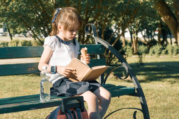 ritratto di studentessa di 7 anni su una panchina che legge libro, mangiando gelato. parco cittadino di sfondo - 6 7 years little girls child portrait foto e immagini stock