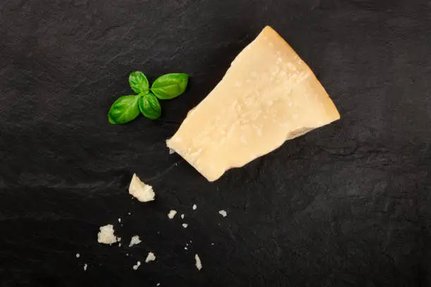 A piece of aged Parmesan cheese with crumbs, shot from the top on a black background with fresh basil leaves and a place for text