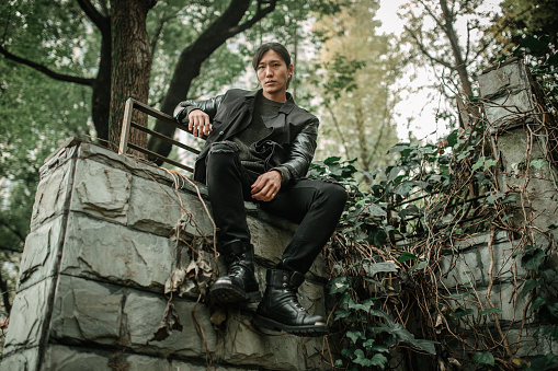 Handsome Asian man with long hair and black outfit sitting on concrete wall