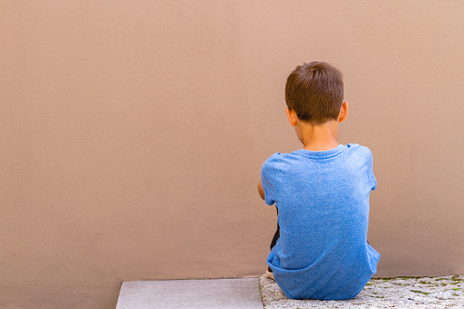 Sad alone boy sitting on the ground behind the wall outdoor.